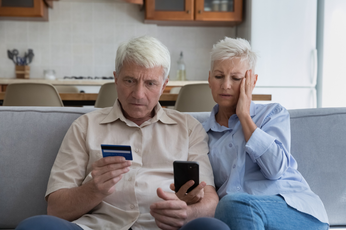 A concerned elderly couple on a couch looking at a cellphone while holding a credit card.