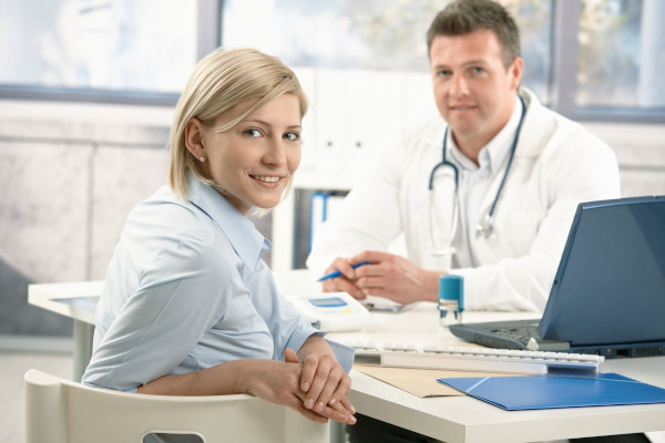 Smiling woman sitting in doctor's office on appointment, looking at camera, doctor in background.